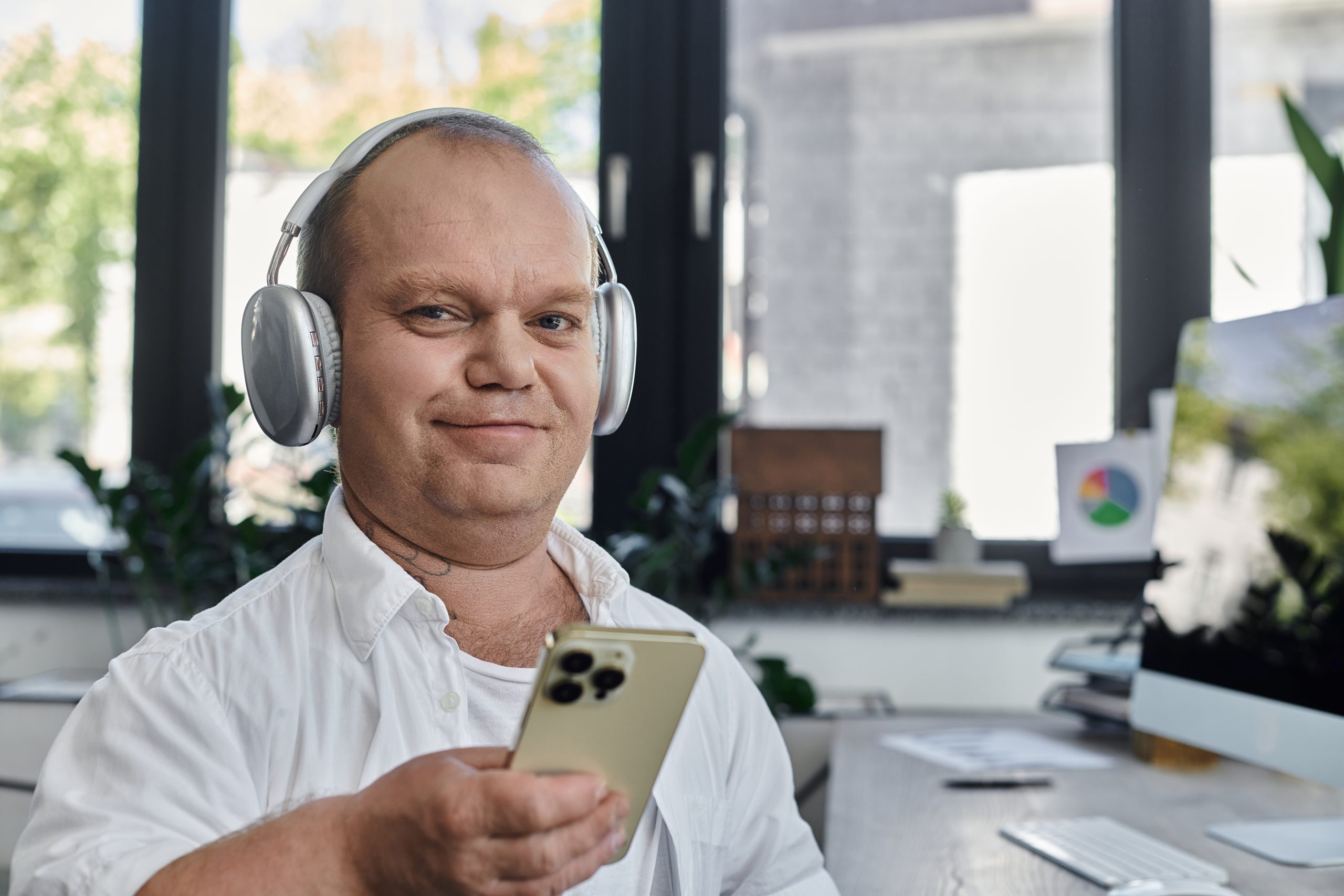 Un homme autiste porte un casque d'écoute et tient un téléphone mobile dans sa main, en souriant et en étant est assis à son bureau dans un décor de bureau